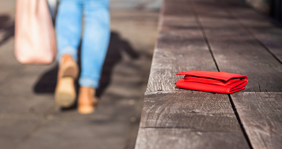 A woman in jeans walks past a red wallet which has been left on a wooden bench in an outdoor public place 